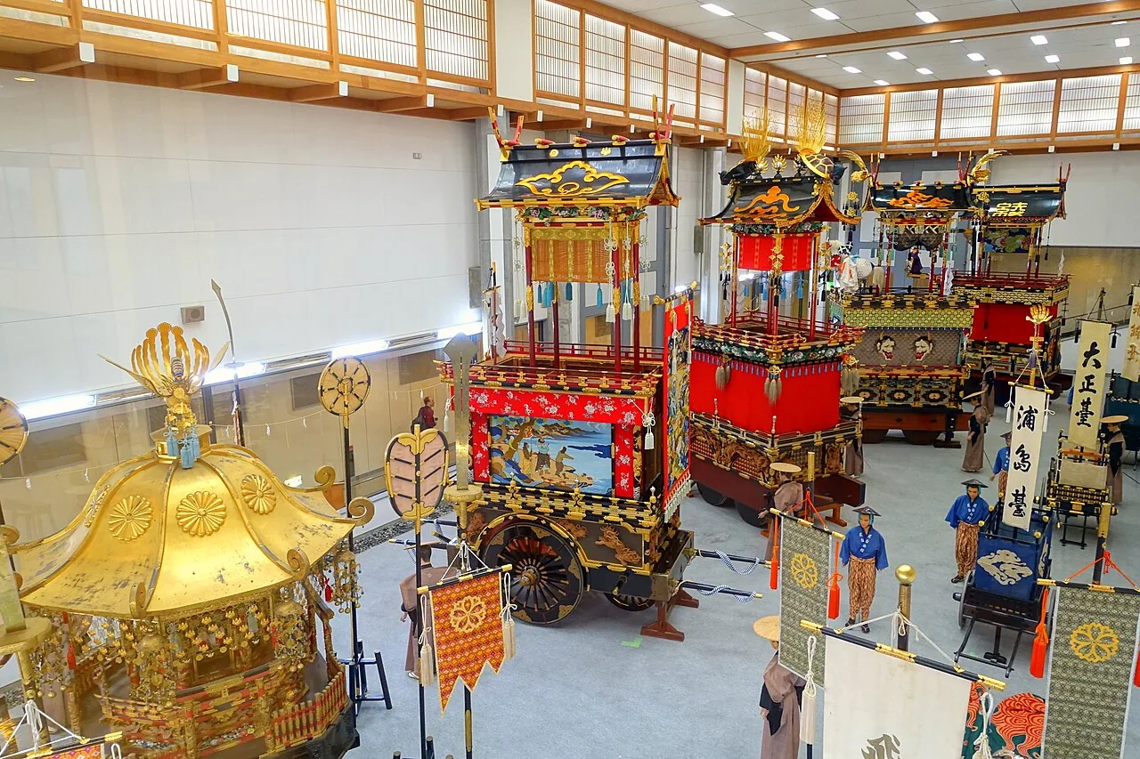 Gilded and red festival floats with costumed figures on display inside a large hall